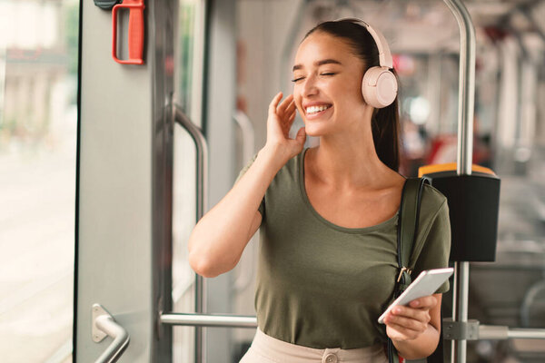 Portrait Of Joyful Passenger Lady Holding Cellphone Wearing Headphones Sitting In Modern Tram Interior, Enjoying Comfortable Public Train Ride And Great Musical Playlist In Transport. City Commute