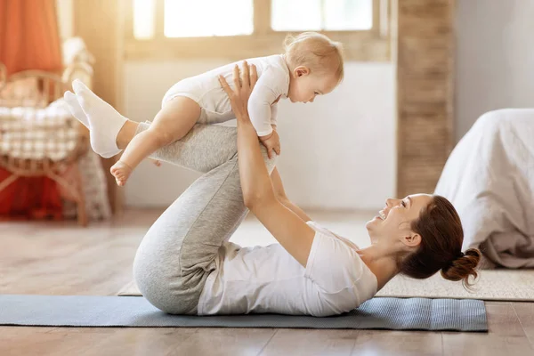 Sport, motherhood and active lifestyle concept. Side view of young mother workout together with her baby toddler in living room at home. Sporty mom having fun and playing with her little infant child