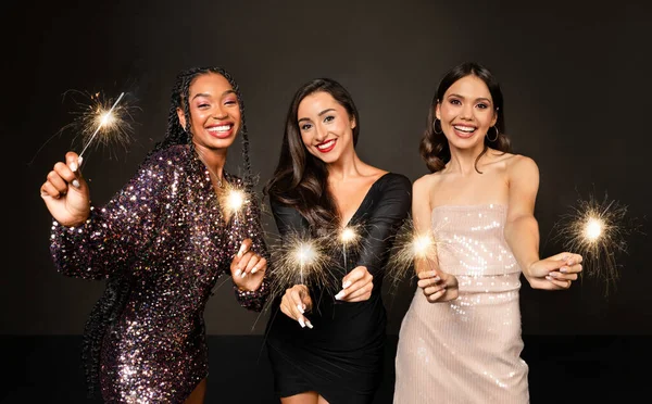 Crazy party time of three beautiful stylish women in elegant outfit celebrating new year 2024 together over black background, young ladies having fun, dancing, holding bengal lights and smiling