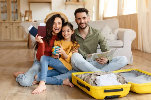 Family Vacation Tour Offer. Joyful Parents And Daughter Sitting With Suitcase, Boarding Pass Tickets And Touristic Map, Celebrating Holiday Journey At Home, Packing Luggage And Planning Trip