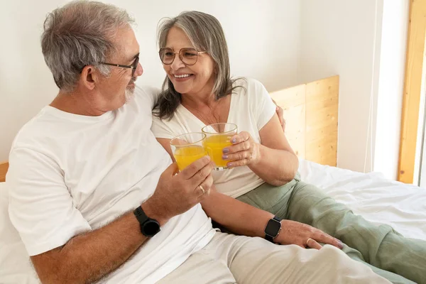 Cheerful old european husband and wife have breakfast, cheers, enjoy glasses of juice, sit on bed in bedroom interior. Love at weekend, rest and relaxation together, health care at home