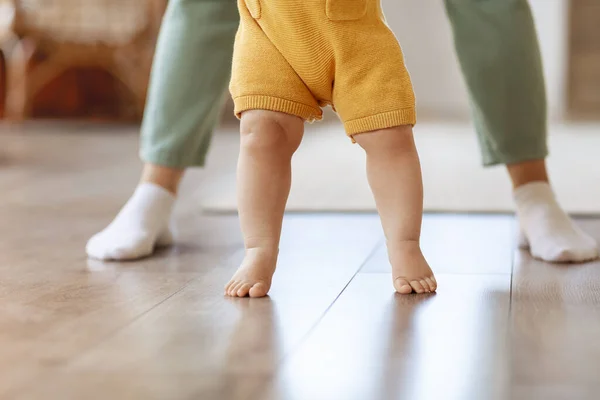 Cropped of caring mom holding small toddler daughter or son hands make first steps together. Legs of little baby kid child learn walking on wooden floor in home with mother. Childhood concept