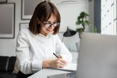 Genç iş kadını masada oturuyor ve Webinar 'a katılırken not alıyor ya da kafede dizüstü bilgisayarla eğitim görüyor. Öğrenci bayan çevrimiçi öğrenim görüyor, eğitime katılıyor.