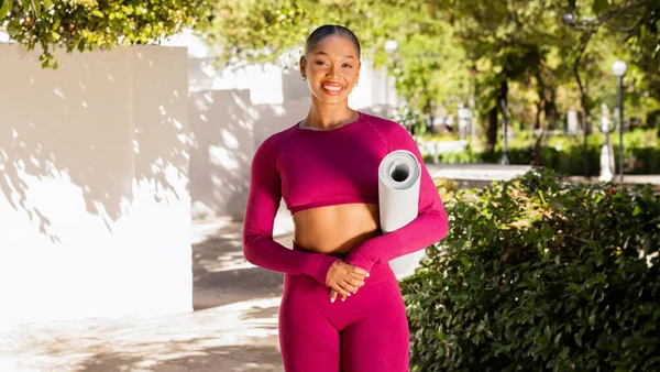 Positive beautiful well-fit athletic young black woman wearing pink tight sportswear posing with fitness mat at garden or park, smiling at camera. Outdoor workout, panorama with copy space