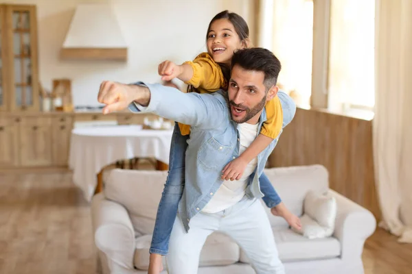 Family Of Super Heroes. Funny Dad Piggybacking His Kid Daughter, Making Superhero Gesture Together, Posing And Having Fun Playing At Modern Home Interior. Joyful Weekend With Father Concept
