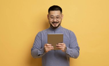 Sarı Stüdyo Arkaplanı 'nın başında dikilirken Dijital Tablet kullanan gülümseyen Asyalı genç adam, Yeni Uygulama Gözlüklü Yakışıklı Adam veya Modern Gadget' ta Alışveriş İnterneti, Kopya Uzay
