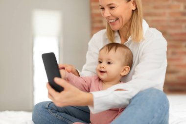 Mutlu Avrupalı anne ve kız bebek cep telefonu kullanarak eğitim oyunları oynayarak ya da çizgi film seyrederek, yatakta oturarak. Bebek ve alet kullanımı kavramı