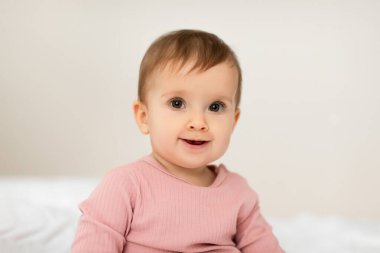 Küçük, sevimli bir kızın portresi. Işıklı yatak odasındaki kameraya bakıp gülümsüyor. Sıradan kıyafetler giyen tatlı bir kız. Bebeklik moda konsepti