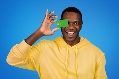 Sweatshirt 'lü neşeli, yakışıklı Afro-Amerikan adam kredi kartını göz göze bastırıyor, eğleniyor, mavi stüdyo arka planında soyutlanıyor. Satış, finans, alışveriş ve para, reklam ve teklif