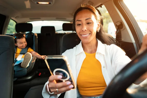 快乐的亚洲妈妈一边开车一边用智能手机 一边和她可爱的小女儿一起在车里安全的座位上开车 有选择地关注电话妇女 母性多重性 — 图库照片