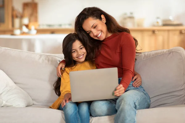快乐的年轻的阿拉伯母亲和女儿使用笔记本电脑 上网冲浪和在线观看卡通片坐在沙发上在家 妈妈和小女孩通过电脑浏览互联网 — 图库照片