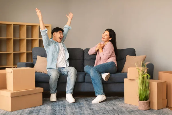 Wow Real Estate Offer. Joyful Korean Couple Celebrating Moving Their Dream Home, Sitting On Sofa And Expressing Joy Among Cardboard Boxes. Family Housing And Relocation Concept
