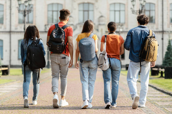 Group of five students with backpacks walking at university campus together, rear view of multiethnic young people going to classes, diverse college friends spending time outdoors after lessons