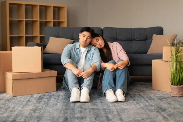 Real Estate Offer. Tired Asian Couple Sitting After Exhausting Relocation, Posing On Floor Among Carton Moving Boxes At New Home Interior. Move House Without Problems And Exhaustion
