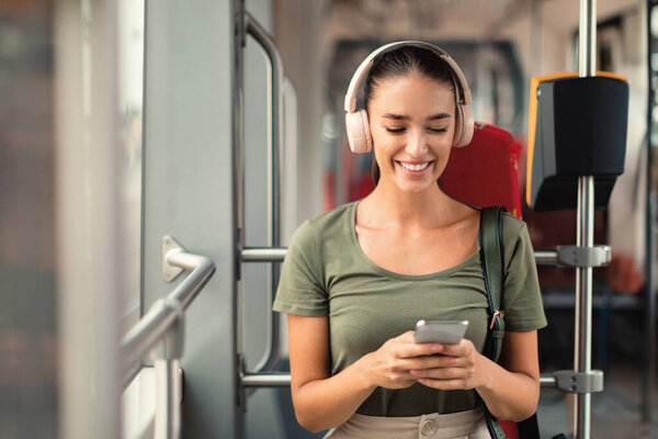 City Commute Soundtrack. Happy Woman Passenger Enjoying Music with Headphones and Smartphone Sitting Comfortably in Modern Tram Inside, Texting While Traveling By Public Transport