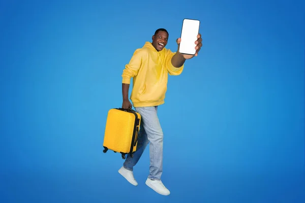 令人高兴的是 年轻的非洲裔美国人带着手提箱走了 在空中冻住了 展示了屏幕空空如也的智能手机 在蓝色的工作室背景下与世隔绝 周末推荐应用程序 — 图库照片