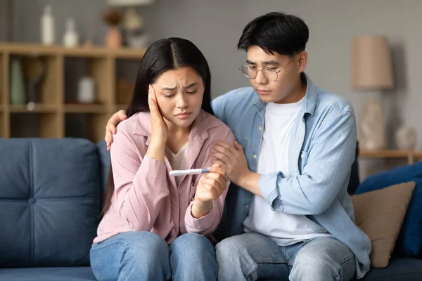 Childbirth Problem. Depressed Korean Couple Holding Negative Pregnancy Test, Sitting Together On Couch At Home. Young Spouses Struggling With Infertility Issue And Dreaming About Baby