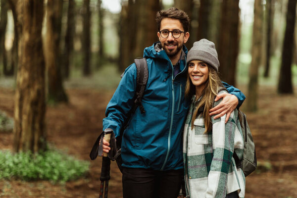 Positive young caucasian couple in jackets with trekking sticks walk in forest together, enjoy trip, active lifestyle, outdoor. Travel hiking, adventure and tourism, relationships
