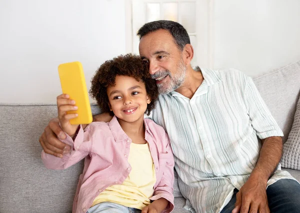多代移动乐趣 快乐的爷爷和小孙子通过智能手机让自己在一起 分享社交媒体应用中的结合时刻 坐在家里的沙发上 — 图库照片