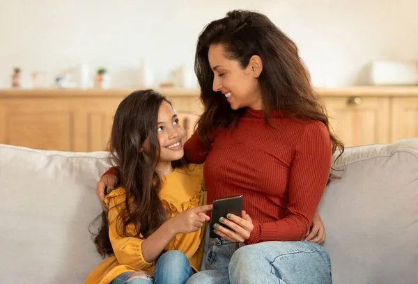 Happy Middle Eastern Mom And Little Daughter Using Smartphone, Kid Girl Pointing Finger At Gadget, Showing Her Favorite Mobile Game Application, Hugging Sitting On Couch At Home Interior