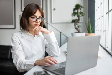 Gözlüklü düşünceli beyaz kadın laptop ekranına bakıyor, iş yerinde oturuyor, problem çözümü hakkında düşünüyor. Masada bilgisayarla çalışan düşünceli bir iş kadını.