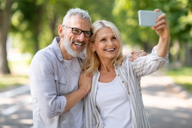Mutlu olgun çift parkta yürürken akıllı telefonuyla selfie çekiyor, neşeli yaşlı eşler dışarıda fotoğraf çekiyor, cep telefonu kameralarına sarılıyor ve gülümsüyor, birlikte vakit geçiriyorlar.