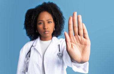 Beyaz önlüklü ciddi bir siyahi bayan doktor mavi stüdyo arka planında izole edilmiş bir şekilde el hareketini durduruyor. Hastalıklara, işe, mesleki tavsiyelere, tıbba karşı mücadele, işaret yok