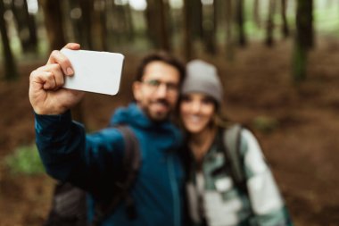 Beyaz ceketli olumlu genç adam ve bayan seyahat tatilinin tadını çıkarıyorlar. Ormanda, açık havada, bulanık bir şekilde akıllı telefondan selfie çekiyorlar. Macera ve turizm blogu, yürüyüş, aktif yaşam tarzı