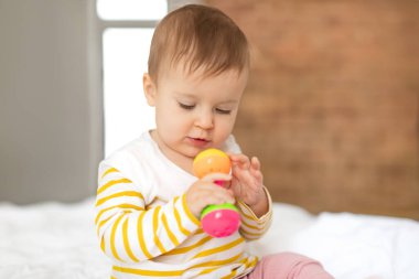 Elinde çıngıraklı oyuncakla elinde bembeyaz çarşaflarla yatak odasında oturan, evde oynayan ve dinlenen tatlı bir kız çocuğu, yakın plan, kopyalama alanı.