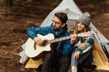 Ormandaki ceketli olumlu beyaz çift çadır, dinlenme, gitar, açık havada gezintilerin tadını çıkarıyor. Müzik, yaşam tarzı, macera ve turizm ile yürüyüş, kamp