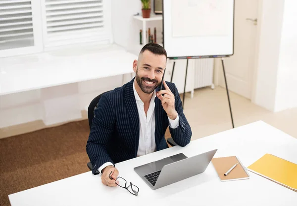 ポジティブな慎重な白人成熟した男は スーツにひげを着て テーブルに座って ラップトップを使用し コワーキングオフィスのインテリアで電話をかけます ミーティング コミュニケーション ビジネスマン ビジネス マネージャーの仕事 — ストック写真