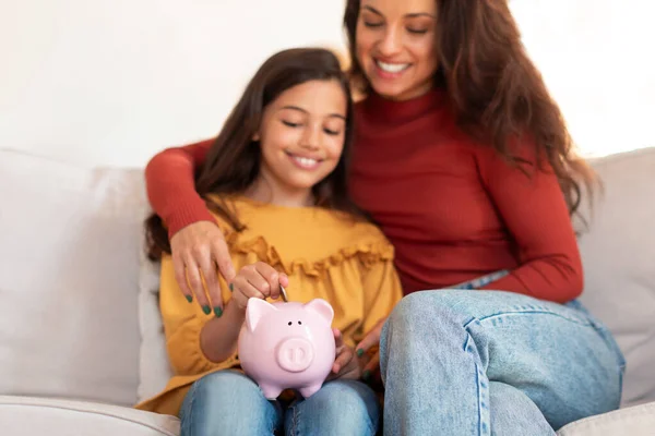 Child Personal Savings. Happy Arabic Mom And Little Daughter Putting Coin In Piggybank, Raising And Saving Money Investments Sitting On Sofa At Home. Finances Safety. Selective Focus On Piggy Bank