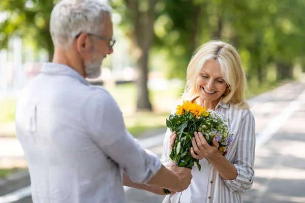Loving Mature Man Giving Flowers To His Wife During Romantic Date Outdoors, Caring Senior Husband Surprising Happy Woman With Bouquet, Greeting With Anniversary Or Birthday While They Walking In Park