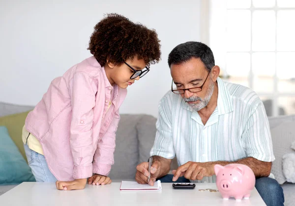 Finance Education. Hispanic Grandfather and Little Grandson Collaborating on Savings, Calculating Money with Calculator, Notes, and Piggybank on Table at Home. Learning Financial Literacy Together