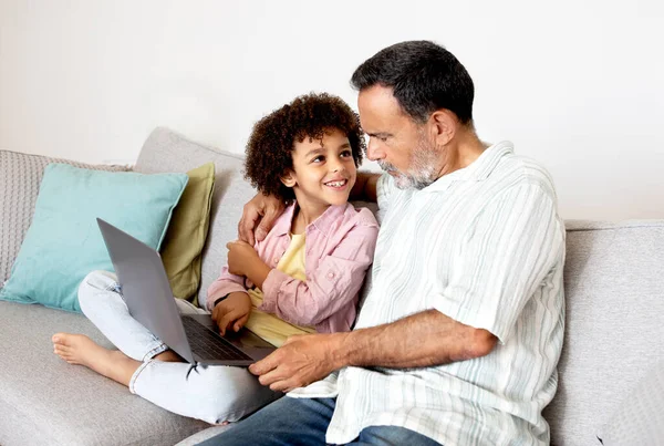 Cheerful Hispanic Senior Grandfather And Little Grandson Using Laptop Hugging On Couch At Home. Tech Savvy Grandparent and Grandchild Embracing Digital Bonding Through Web, Browsing On Computer