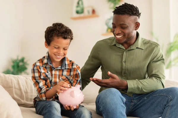 Kids Pocket Money And Personal Savings. Happy Son And His African American Dad Putting Coin In Piggybank Sitting On Sofa In Modern Living Room At Home. Bank Offer, Financial Investments Safety