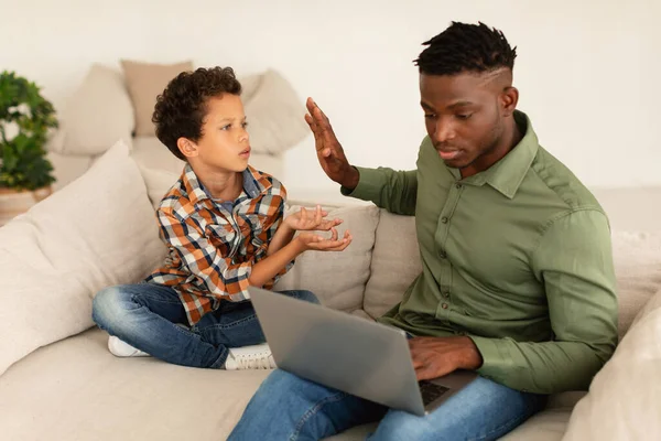 Indifferent Father. Busy African American Dad Ignoring His Son, Using Laptop Working Online On Computer And Gesturing Stop To Kid Boy Sitting On Sofa At Home. Daddy And Child Relationship Issue