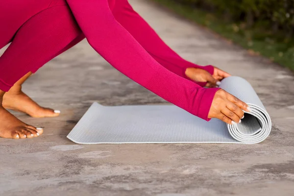 Cropped of black woman in pink sportswear unrolling fitness mat before yoga practice outdoor. Lady athlete getting ready for pilates training at park or garden, copy space