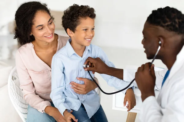 Child Health Checkup. Mother And Son Visiting Family Doctor, Black Pediatrician Man Using Stethoscope Listening To Kid Boys Heartbeat Via Stethoscope, During Medical Appointment In Clinic Indoor