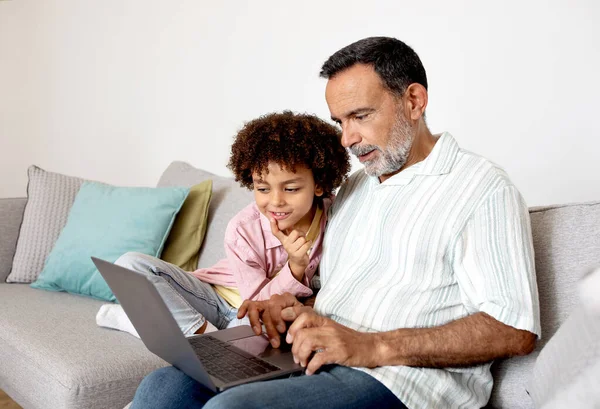 Hispanic Grandpa And Little Grandson Using Laptop For Online Entertainment, Watching Film Together At Home. Grandparent And Grandchild Browsing Web On Computer. Generations United Through Gadgets