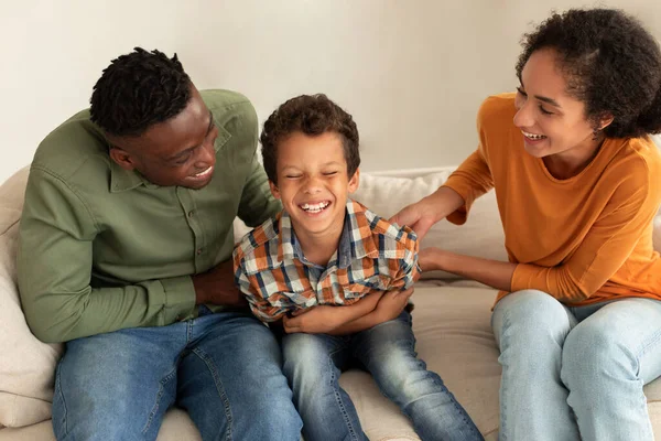 Family Fun. Joyful Diverse Parents Tickling Their Little Son And Laughing Together At Home, Enjoying Playtime, Cuddling And Bonding On Weekend, Sitting On Couch In Cozy Modern Interior