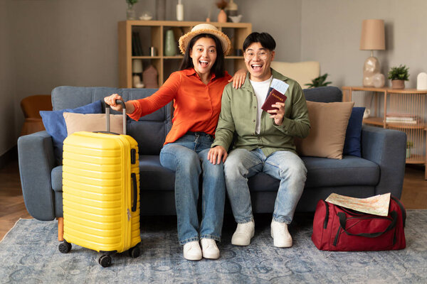 Joyful Japanese Travelers Couple Posing With Travel Tickets And Packed Suitcase, Ready For Summer Trip, Sitting On Sofa At Home, Smiling To Camera. Spouses Tourists Celebrating Vacation