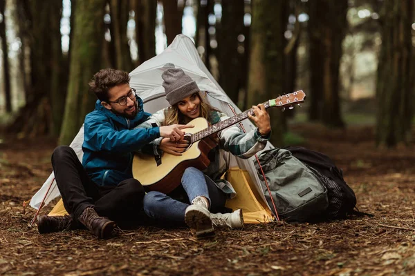 Glad young caucasian guy help lady with music lesson in forest, enjoy travel vacation with tent, playing guitar, outdoor. Camping, hiking, lifestyle, adventure and tourism together