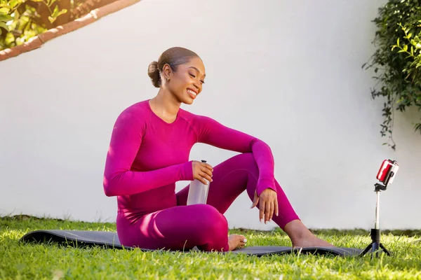 Happy athletic young black woman in pink tight sportswear sitting on fitness mat, drinking water after workout outdoor at garden, look at smartphone screen. Online yoga pilates class, fitness video