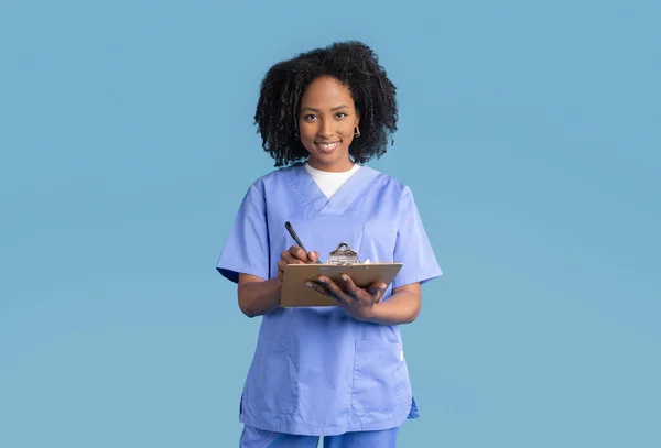 Glad black millennial lady nurse in uniform making notes, isolated on blue studio background. Exam in clinic, work, professional occupation, medicine and health care, consultation, support