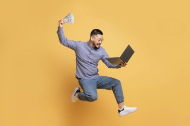 Büyük Kâr. Asyalı heyecanlı genç adam dizüstü bilgisayarla ve sarı arka planda bir dolarla atlıyor, mutlu milenyum adamı, başarılı yatırımını kutluyor ya da internetten piyango kazanıyor, kopyalanıyor.