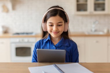 E-Öğrenme Teklifi. Kablosuz kulaklıklı şirin bir kızın portresi dijital tablet kullanıyor, evde otururken ödev yapıyor, internette sörf yapan liseli kız sekme bilgisayarında web sörfü yapıyor.