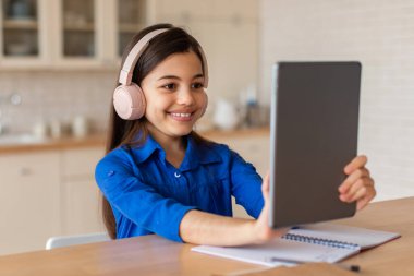 Kablosuz kulaklık takan, dijital tablet bilgisayarla oturan, Modern Ev İçi 'nde rahatlayan ve öğrenen mutlu Arap Öğrenci Kız. Okul çocuğu internette ödev yapıyor.