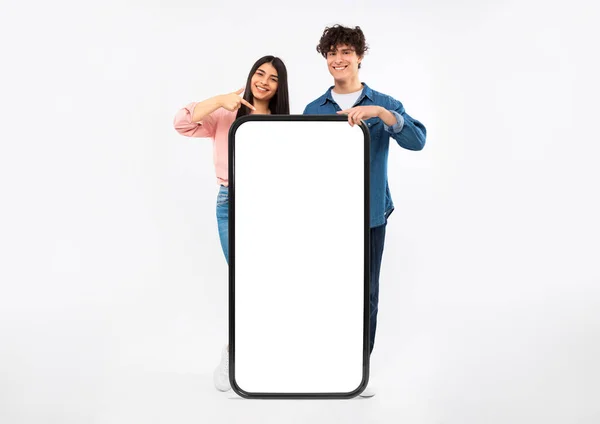 伟大的移动应用程序 快乐的年轻夫妇展示巨无霸智能手机 手指指向空白屏幕为应用程序广告 站在大手机后面的白色工作室背景 莫科普 — 图库照片
