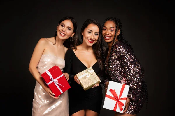 Christmas Gift. Happy multiethnic beautiful young women holding present boxes posing on black studio background. Winter holidays celebration, New Year Celebration and Xmas Gifts concept